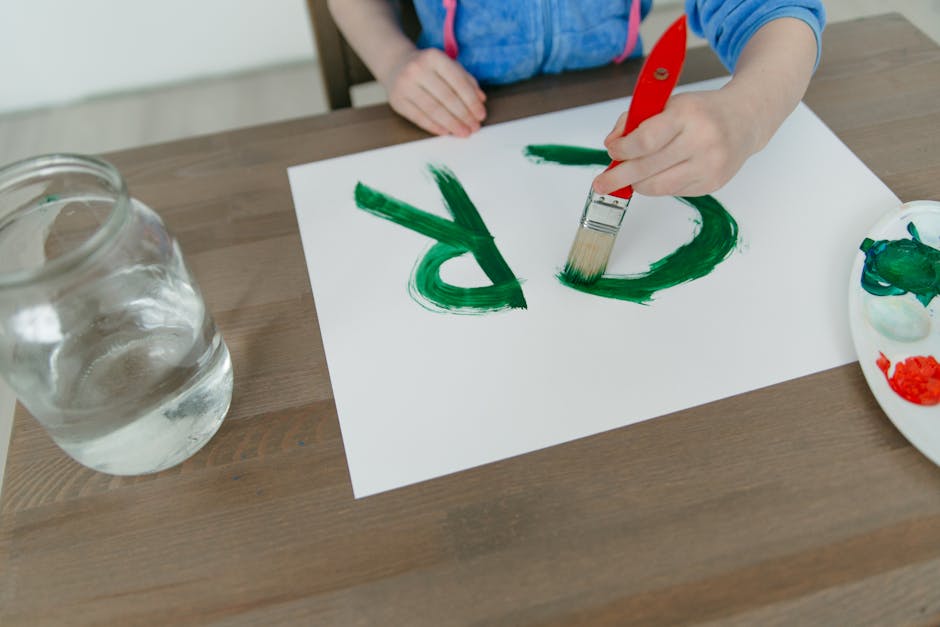 Child painting green letters on paper with a paintbrush. Creative indoor activity captured in detail.